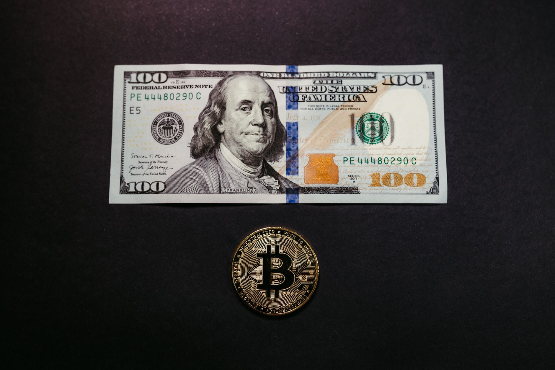 Image of a US 100 dollar bill and a Bitcoin coin on a black background, symbolizing traditional and digital currency.