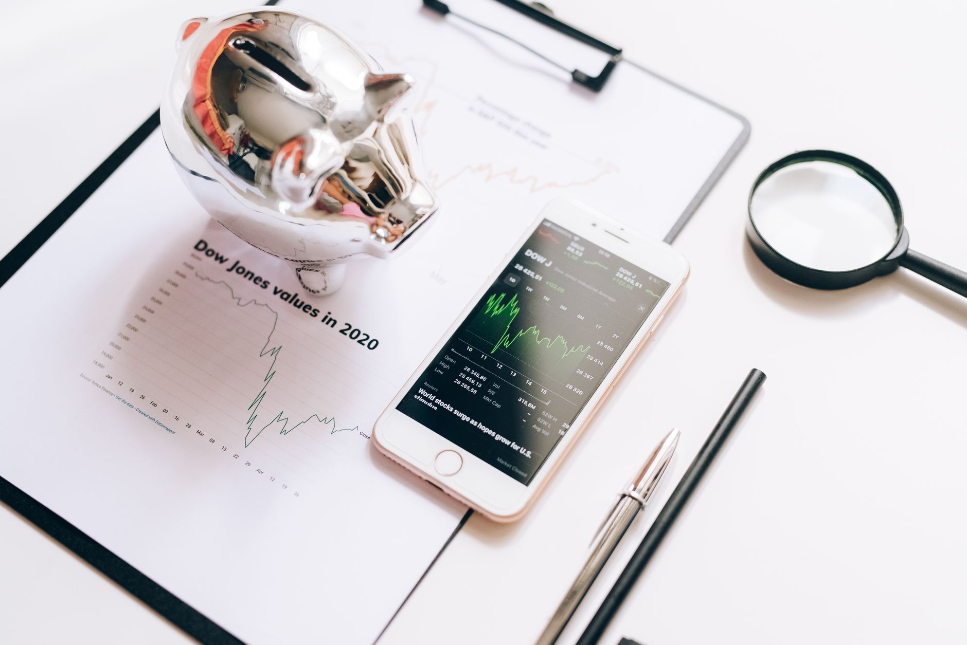 Flat lay of financial tools: chart, smartphone, magnifying glass, and piggy bank.